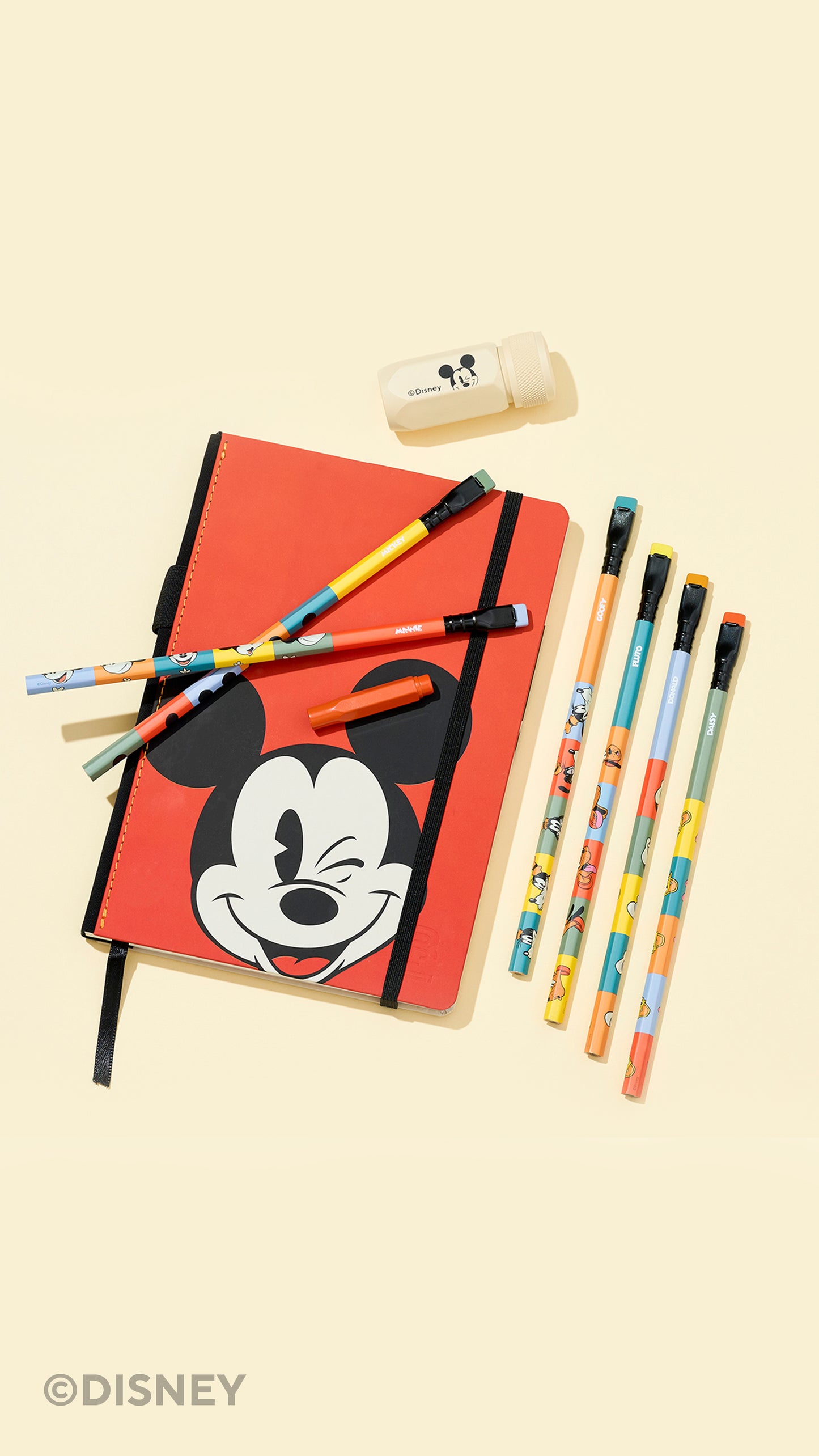 A Disney-themed stationery flat lay featuring a red notebook with a large Mickey Mouse face, several colorful pencils, a small eraser, and a pen on a pale cream background. A Disney copyright mark is visible in the bottom-left corner.